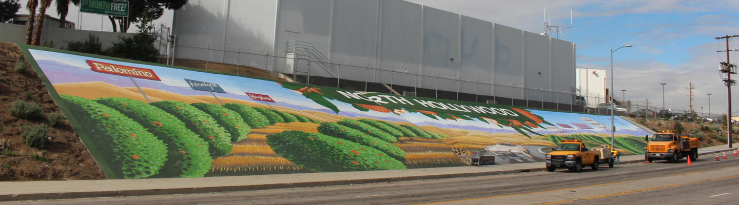 A photograph of the large mural taken from across the street. The mural reveals rows of orange groves with the words “North Hollywood” at the top. Two yellow city trucks are parked in front of the mural.