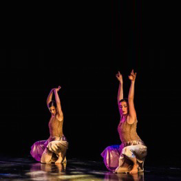 Two dancers squat and raise their hands during a performance on a theater stage.