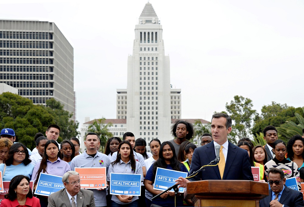 Various participants of the Hire LA Youth program gather behind Mayor Eric Garcetti as he gives a speech at Grand Park. City Hall rises in the background. 
