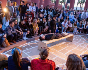 A crowd gathers around DCA Cultural Trailblazer Jay Carlon while they lay on a on a bench during a performance piece at Cal State Los Angeles.