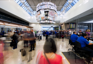 Partnered Arts Program between Los Angeles World Airports and DCA. An architectural photograph of an airport with an artwork installed on the ceiling. Its form resembles a mix of a chandelier and a flight announcement teleprompter. Underneath people move around trying to get to their terminals