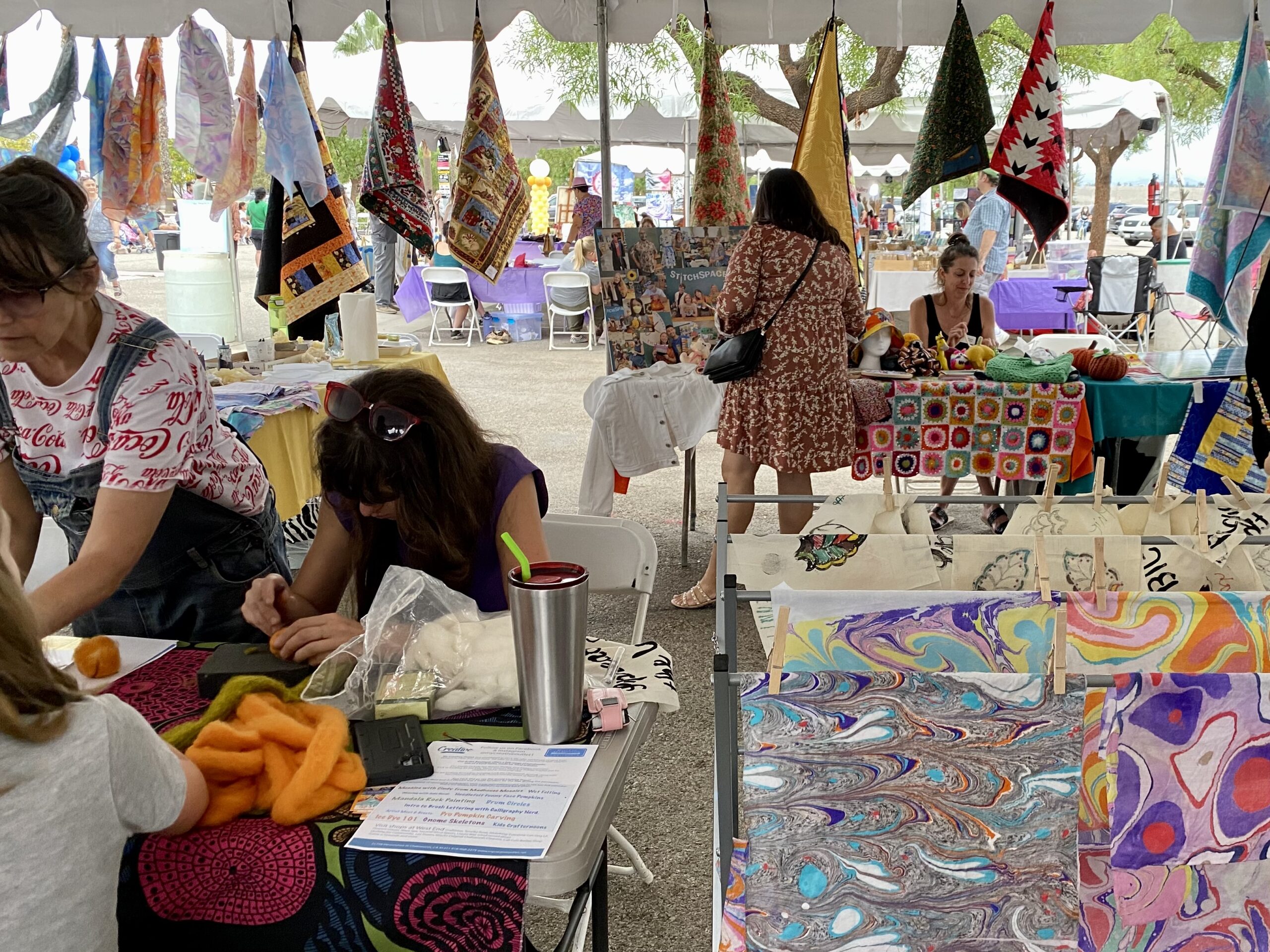 The image shows a group of people sitting at a table with flags. The accompanying text seems to be a list of creative activities or workshops, including topics like mandala rock painting, brush lettering, pumpkin carving, and kids' crafting. The image and text suggest that the event may be an outdoor marketplace or flea market featuring art and creative activities.
